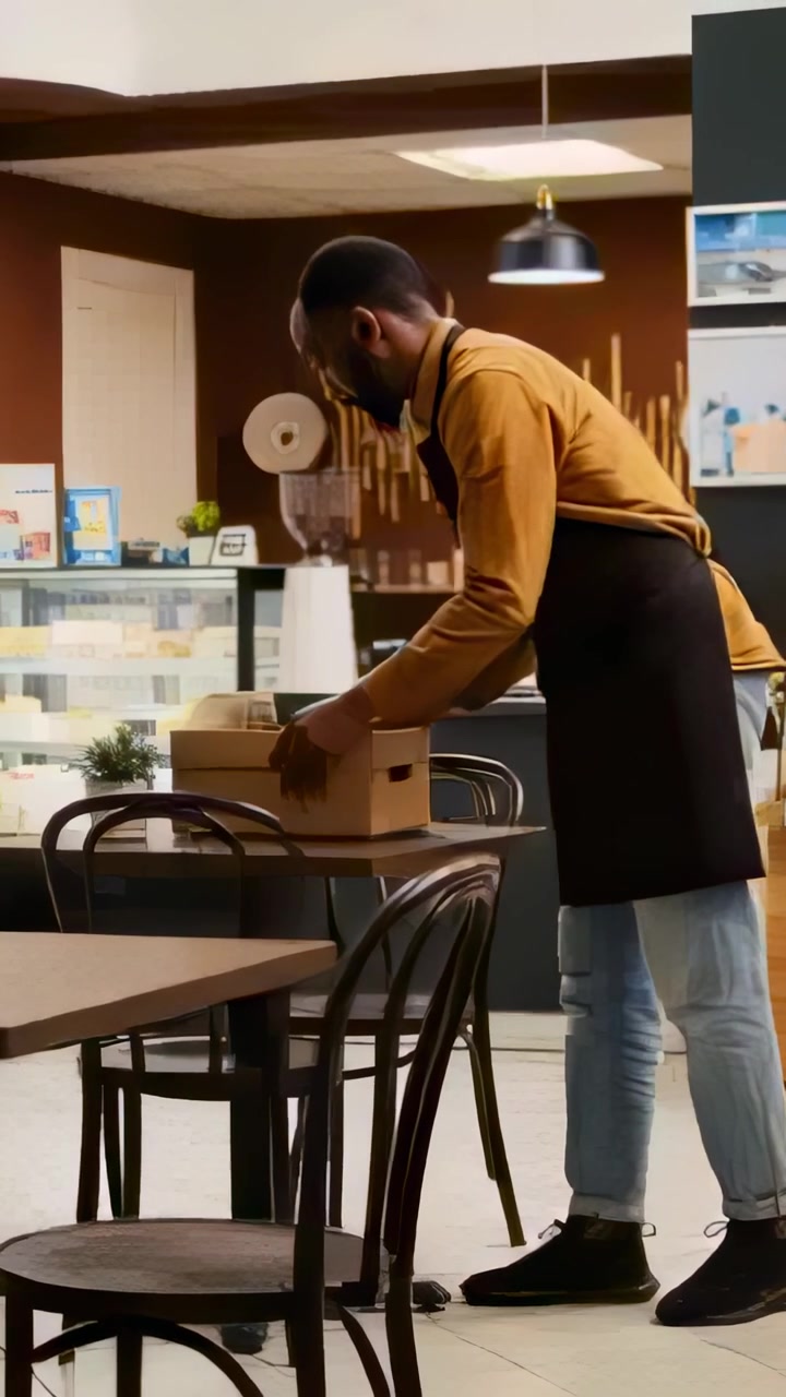 Vídeo vertical: Colocando caixas empilhadas para viagem, barista com blusa mostarda e avental preto arrumando em mesa de café