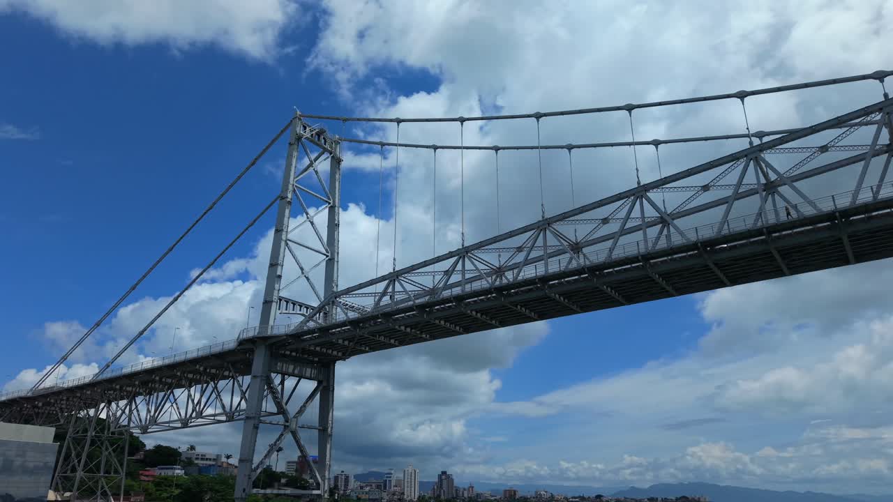 Low angle parallax aerial shot of Hercílio Luz Bridge in Florianópolis Santa Catarina Brazil showing historic steel suspension structure against dramatic blue sky and clouds over the bay
