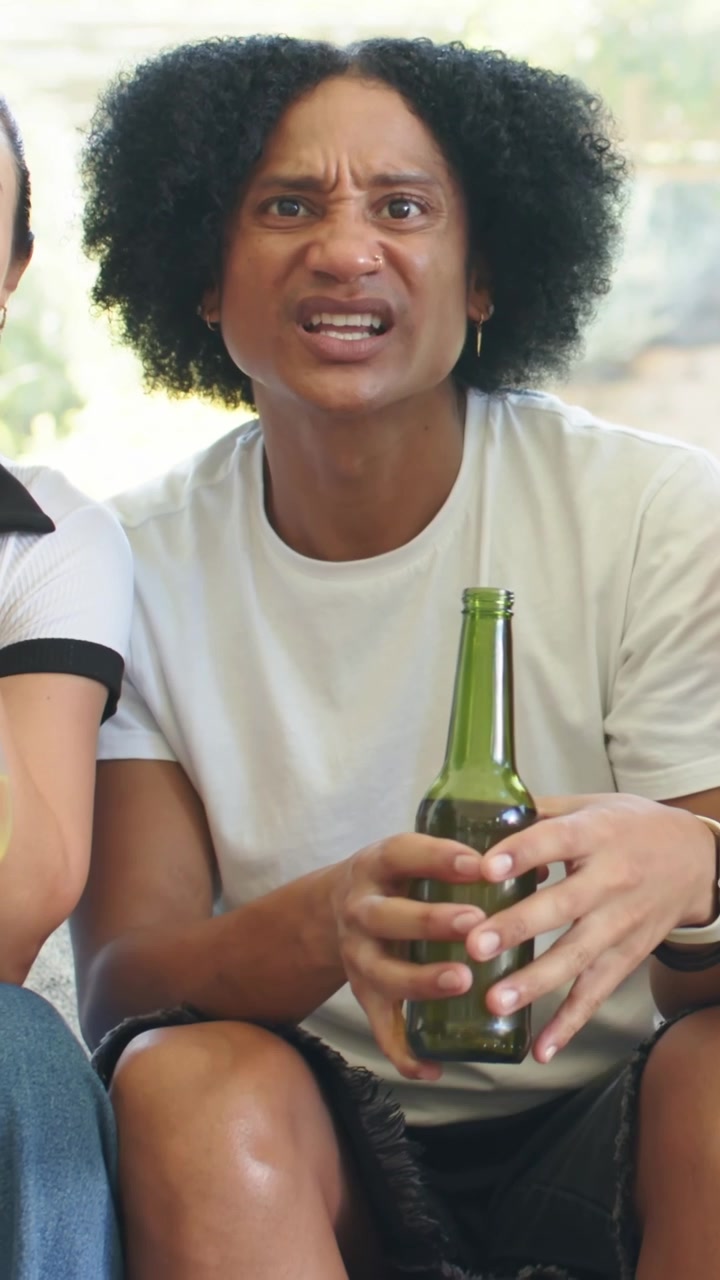 Vertikales Video: Mann im T-Shirt reagiert auf ein Stichwort von außerhalb des Bildes, wird wütend auf dem Sofa mit grüner Flasche.