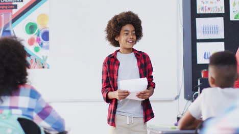 diverse female teacher and happy schoolchildren at desks reciting in school classroom
