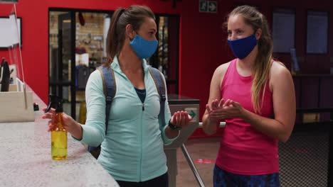 dos mujeres caucásicas con máscaras faciales hablando en el vestíbulo del gimnasio y desinfectando las manos