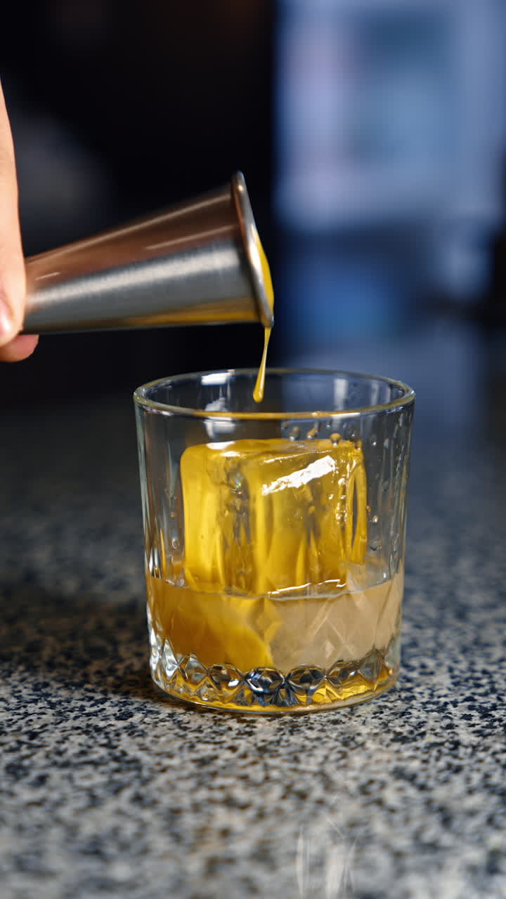 Yellow thick liquid in poured into the glass with ice cube in it. Preparation of alcohol beverage in the bar. Close up. Vertical video.