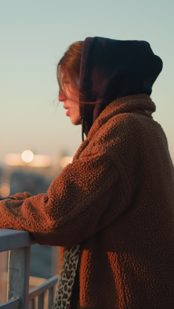 A sorrowful woman walks slowly while holding onto a metal railing, dressed in a brown coat and a hoodie, with a melancholic expression as the cityscape blurs in the background during sunset