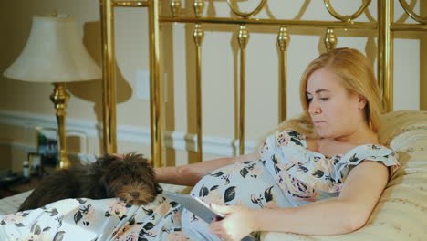 a pregnant woman uses a tablet in her bedroom her dog lies beside her