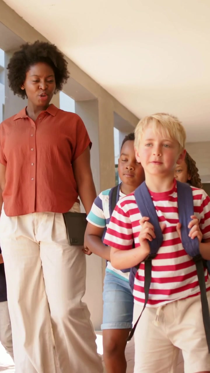 Vertical video: Walking woman in rust shirt chaperoning children along school hall with backpacks