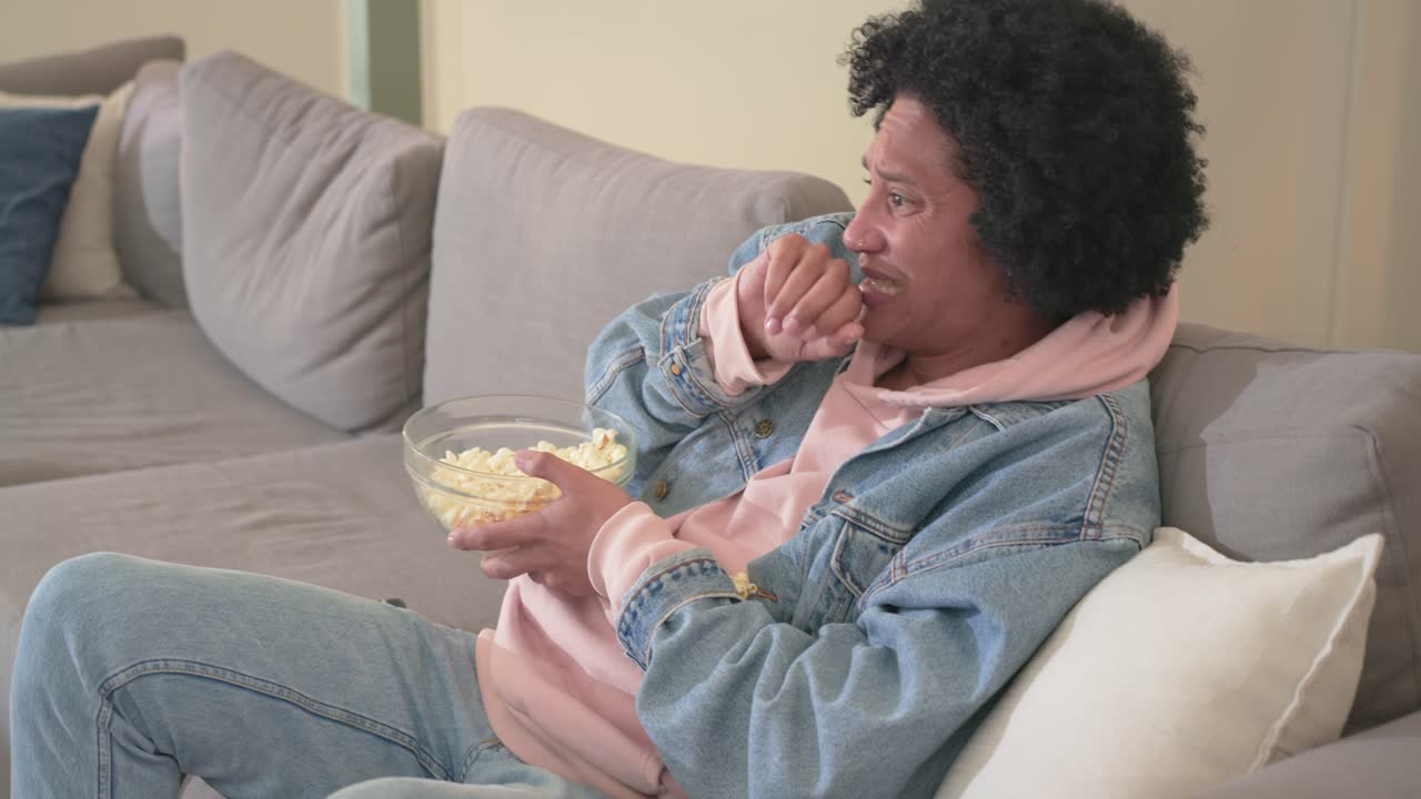 Uomo di mezza età che reagisce a una scena di un film, mangia popcorn e afferra il telecomando sul divano, spazio per il testo