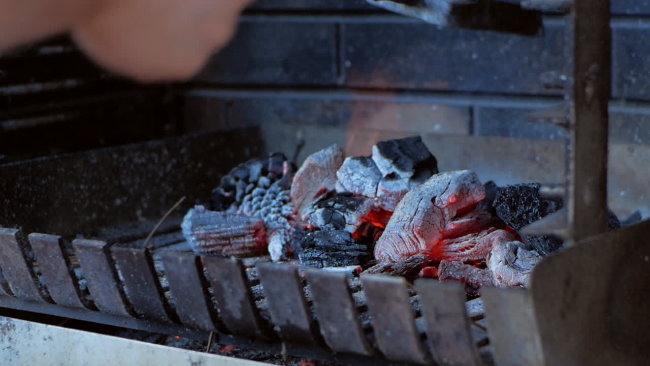 Carboni ardenti e braci roventi bruciano sul barbecue, preparati da una mano caucasica con un attrezzo da camino, primo piano a mano libera.