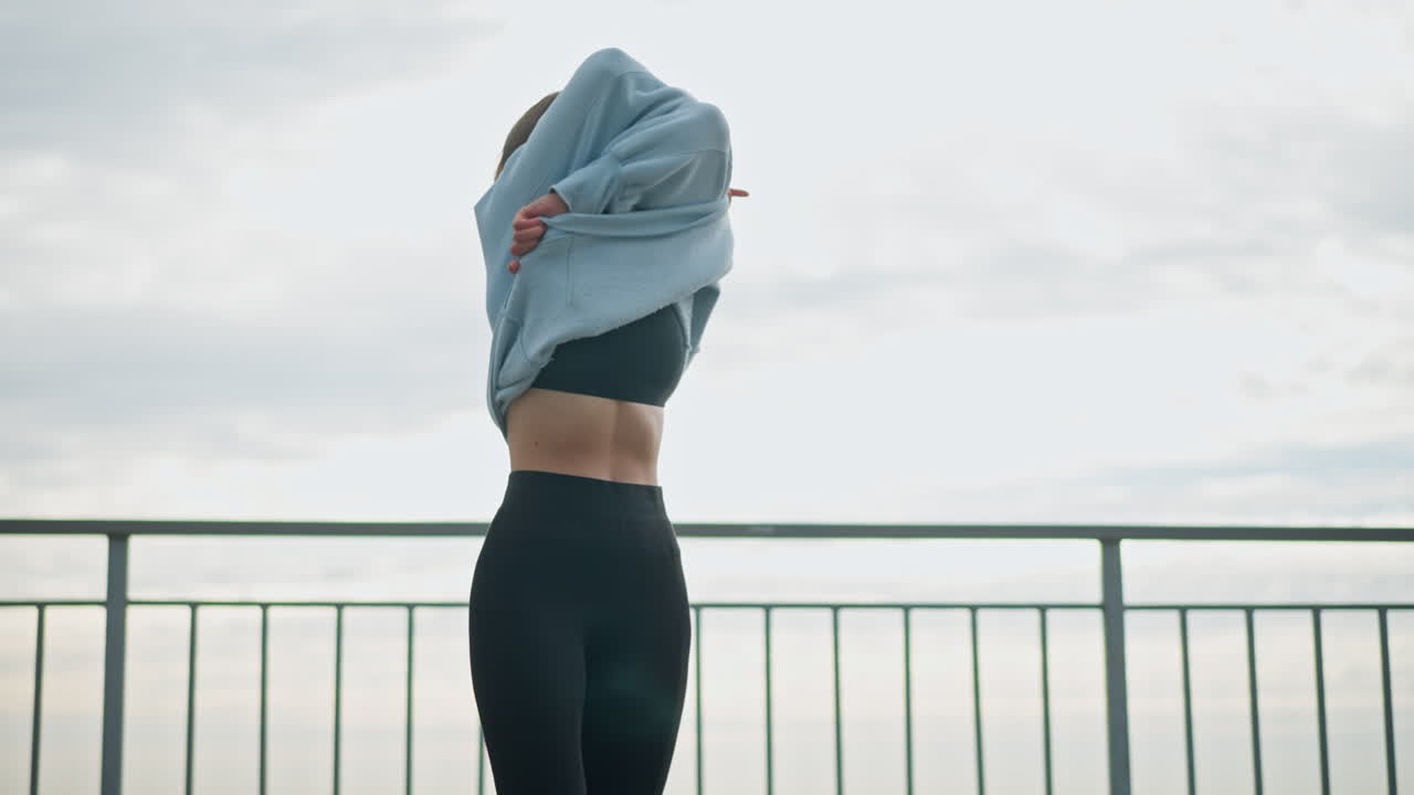 Woman standing beside iron fence, removing hoodie with a smile on her face, she is in an outdoor setting with a calm expression, perfect for fitness, lifestyle, and outdoor moments