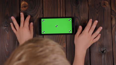 child using a smartphone with a green screen mockup on a wooden background.