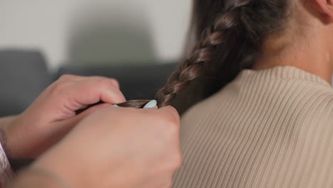 nanny braiding girl’s hair in cozy home, intimate caregiving scene with focused hands weaving strands, child sitting relaxed on sofa, soft natural light, tender personal grooming, peaceful family bond moment