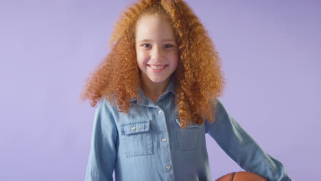 studio shot of young girl holding basketball under arm against purple background