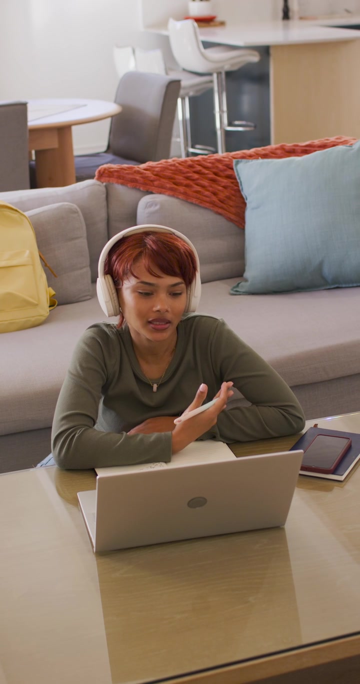 Vertical video: African American woman joining call at laptop, taking study notes at low table