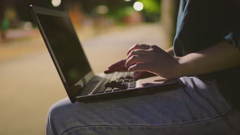 close-up of woman using laptop outdoors at night, fingers typing on keyboard with white reflection on screen, with soft blur background lit up