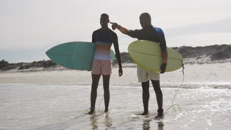 Afroamerikanischer-Vater-Und-Sohn-Im-Teenageralter-Stehen-Am-Strand,-Halten-Surfbretter-In-Der-Hand-Und-Reden