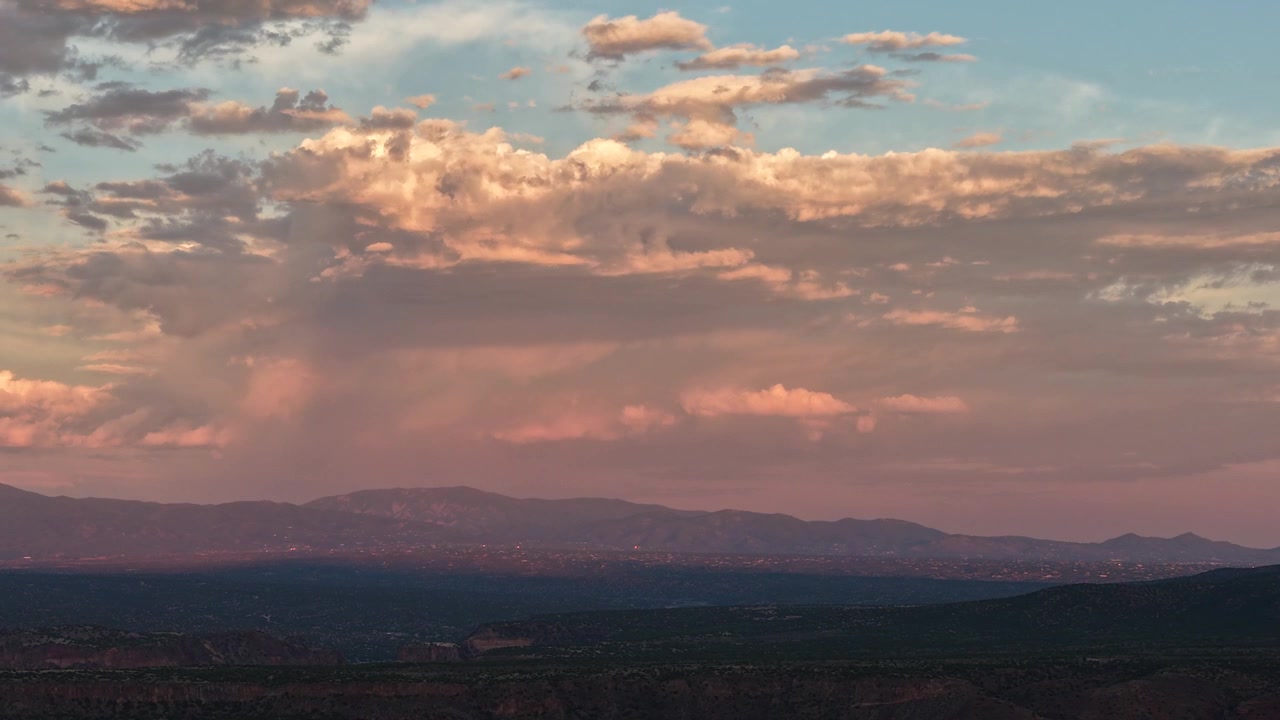 4K drone timelapse of a wave of warm sunset light rippling across majestic, fluffy clouds. The fading evening sun casts a pink glow over the vast landscape and mountains of Santa Fe County.