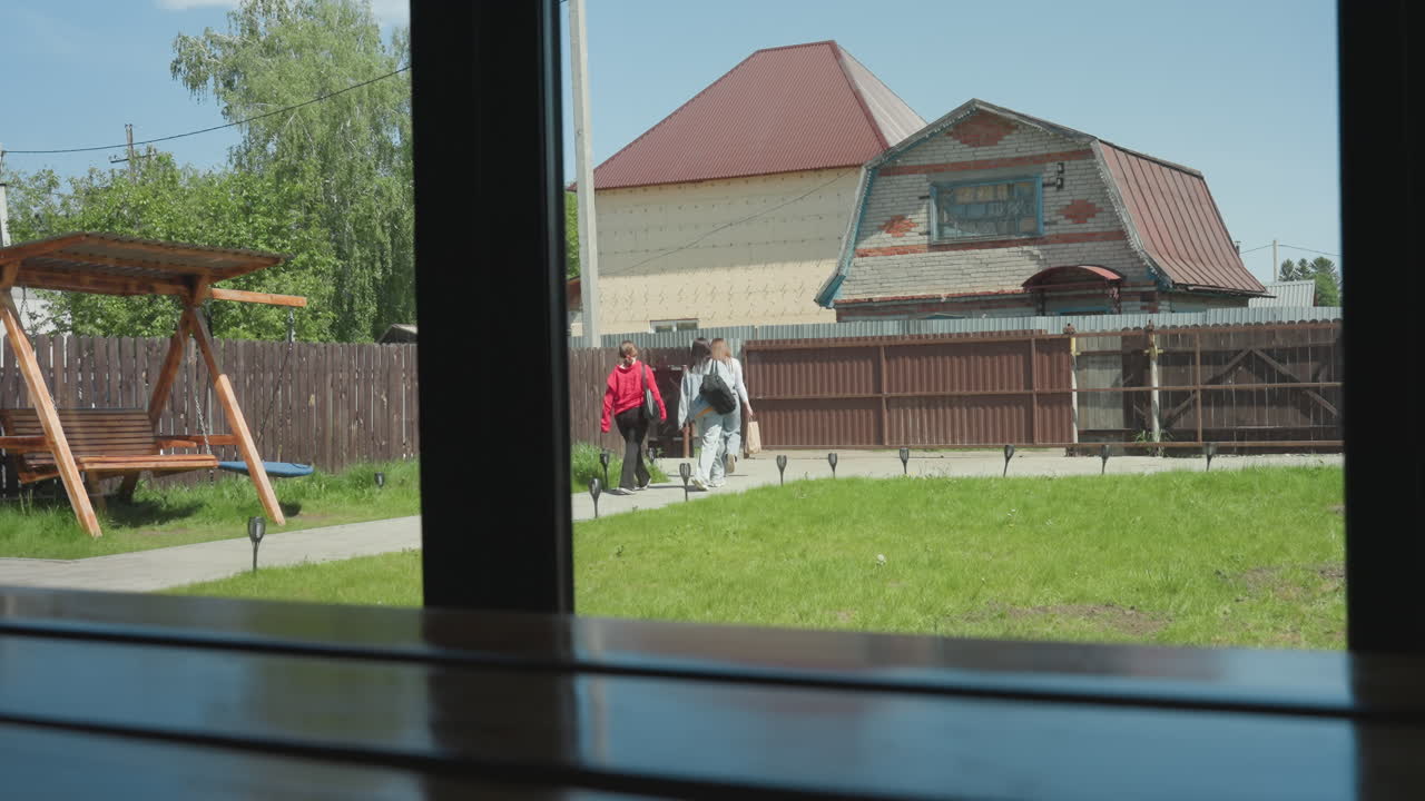 Three ladies seen through glass window walking away from camera on concrete path toward compound gate, with wooden swing, green lawn, and residential houses in background under bright sunny sky