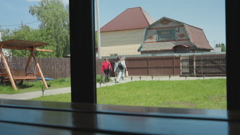 three ladies seen through glass window walking away from camera on concrete path toward compound gate, with wooden swing, green lawn, and residential houses in background under bright sunny sky