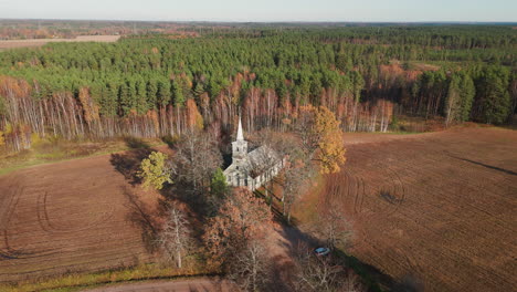 drone captures aerial view of little white ancient church surrounded by empty green fields and forest in autumn beauty