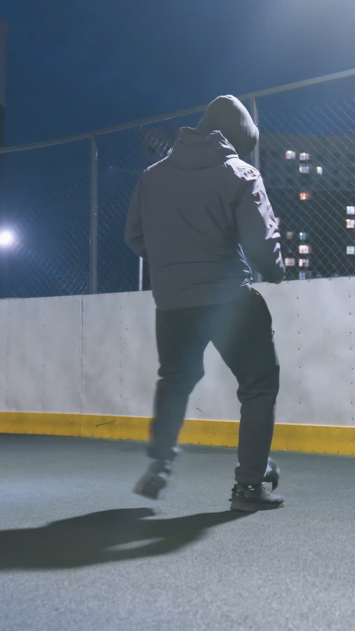 Coach kicking soccer ball against wall on illuminated urban sports field at night, with reflection visible on wall and cityscape lights in background