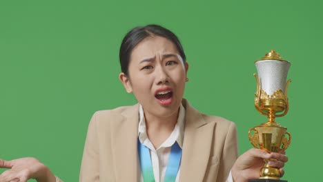 황금 메달을 가진 슈트를 입은 아시아 사업가 여성이 스튜디오의 녹색 스크린 배경에서 트로피를 의심스럽게보고 있습니다.
