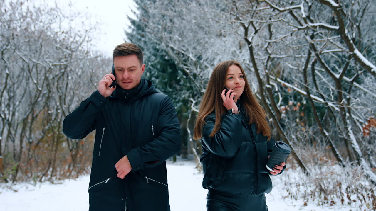Man and woman wearing black jackets walk by the winter nature. People go and speak on the phones.