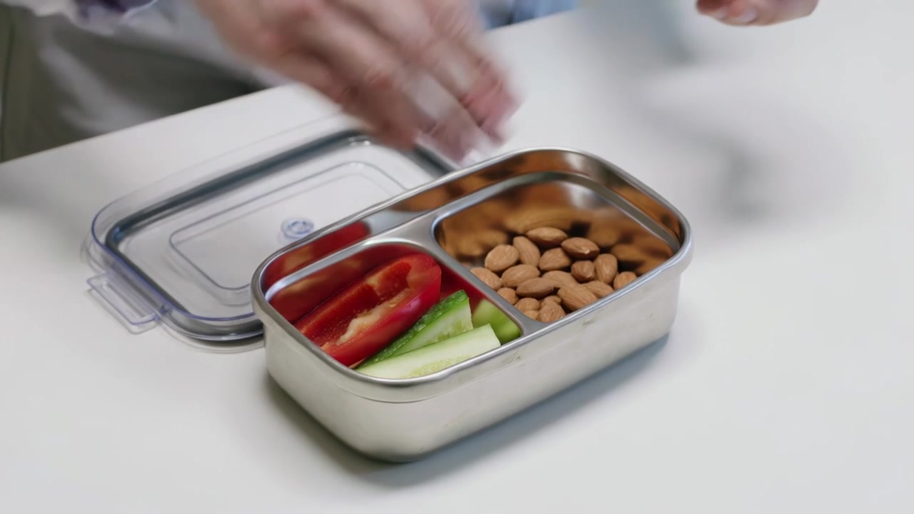 Alcanzando a un hombre con camisa gris preparando el almuerzo en una mesa, añadiendo pepino y pimiento rojo en una lata, compactando almendras.