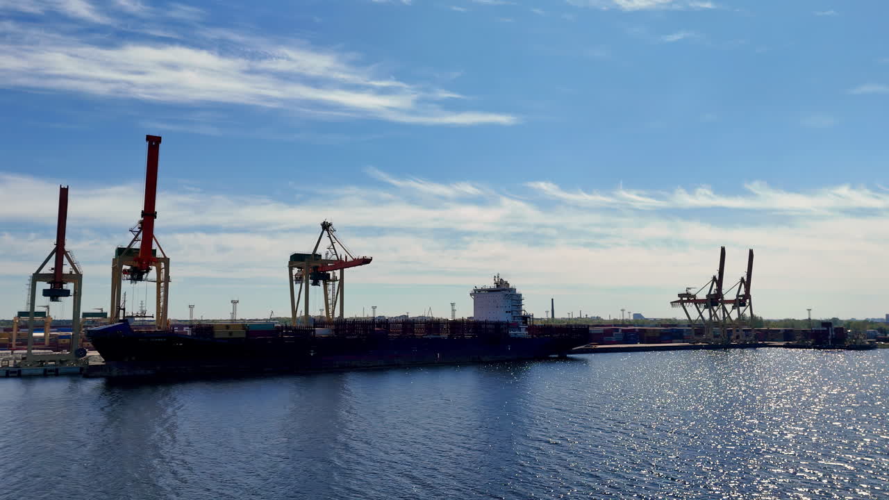 Huge container vessel rests at a busy shipping port with towering cranes positioned for operations, while sunlight reflects across the rippling water under a clear sky