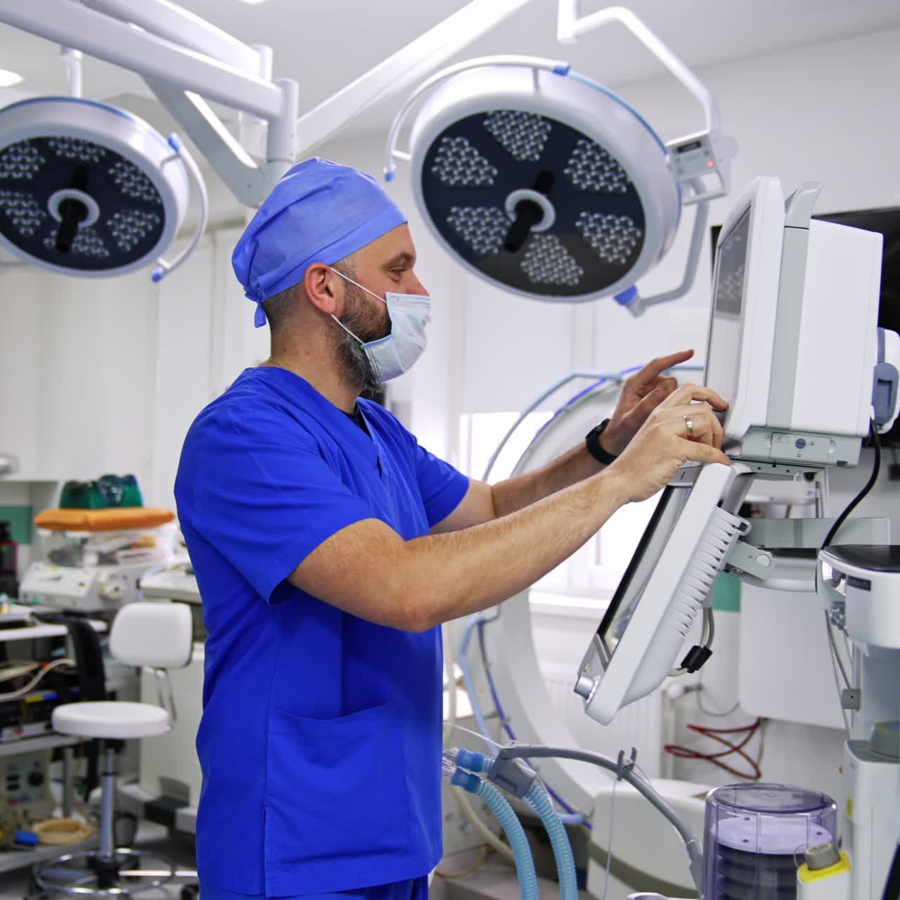 Bearded medic in mask standing near the equipment in surgery room. Anesthesiologist switching on the screens on lung ventilator