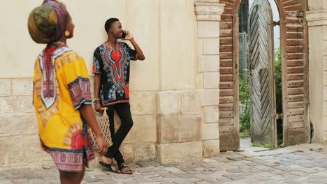 Young-Good-Looking-And-Joyful-Man-In-The-Traditional-Shirt-Speaking-On-The-Phone-Outdoor-In-The-Yard-While-Beautiful-Woman-Passing-By-Him