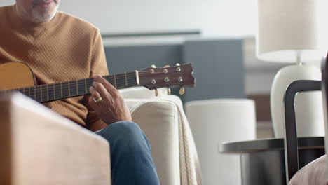 midsection of senior biracial man sitting on couch playing guitar at home, copy space, slow motion