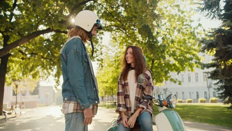 Glückliches-Paar:-Ein-Typ-Mit-Lockigem-Haar-In-Einer-Jeansjacke-Und-Einem-Weißen-Helm-Kommuniziert-Mit-Seiner-Brünetten-Freundin-In-Einem-Karierten-Hemd-Und-Einem-Weißen-T-Shirt,-Die-Auf-Einem-Grünen-Moped-In-Einem-Sommerlichen-Stadtpark-Sitzt