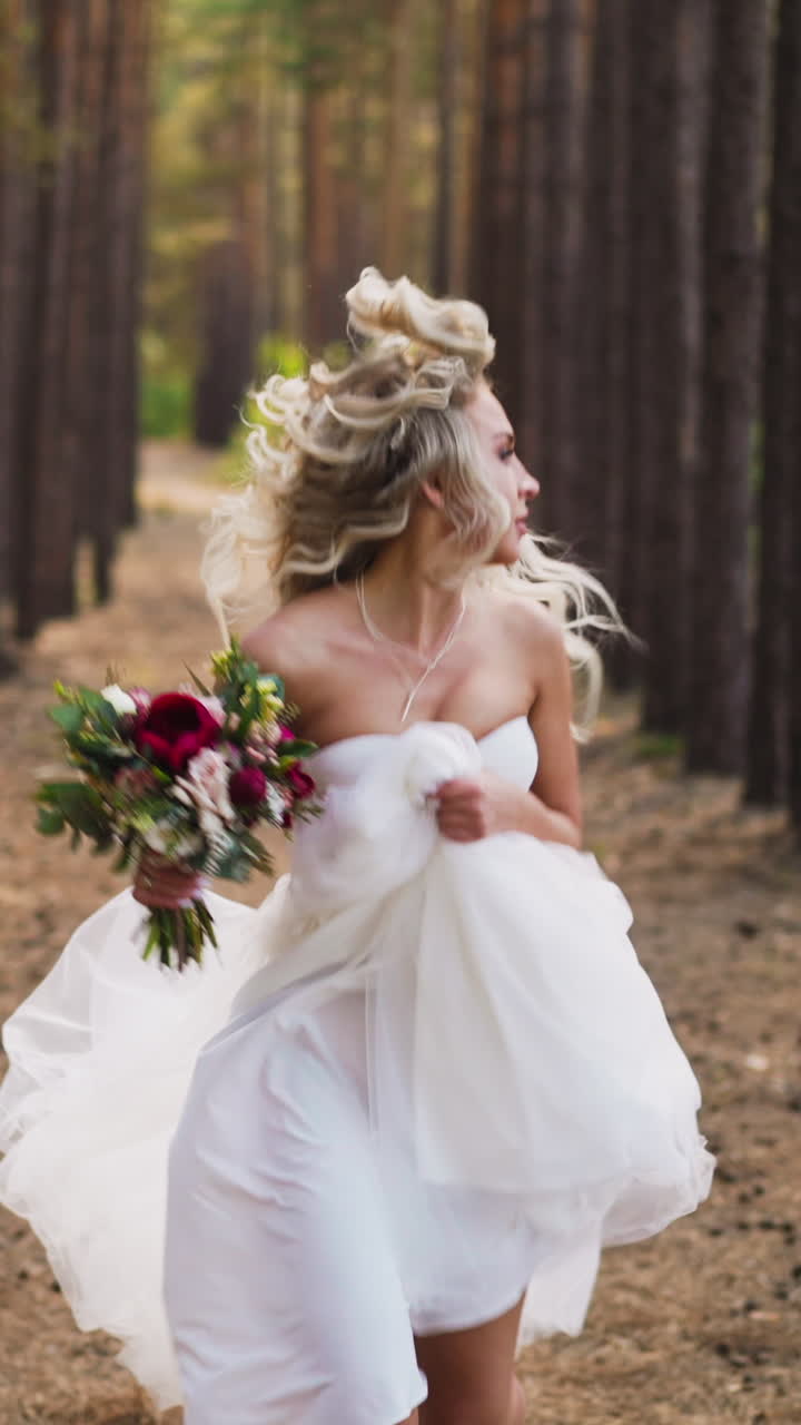 Pretty woman bride in chiffon wedding dress with festive flowers bouquet runs looking back supporting skirt hem along pine forest on summer day slow motion