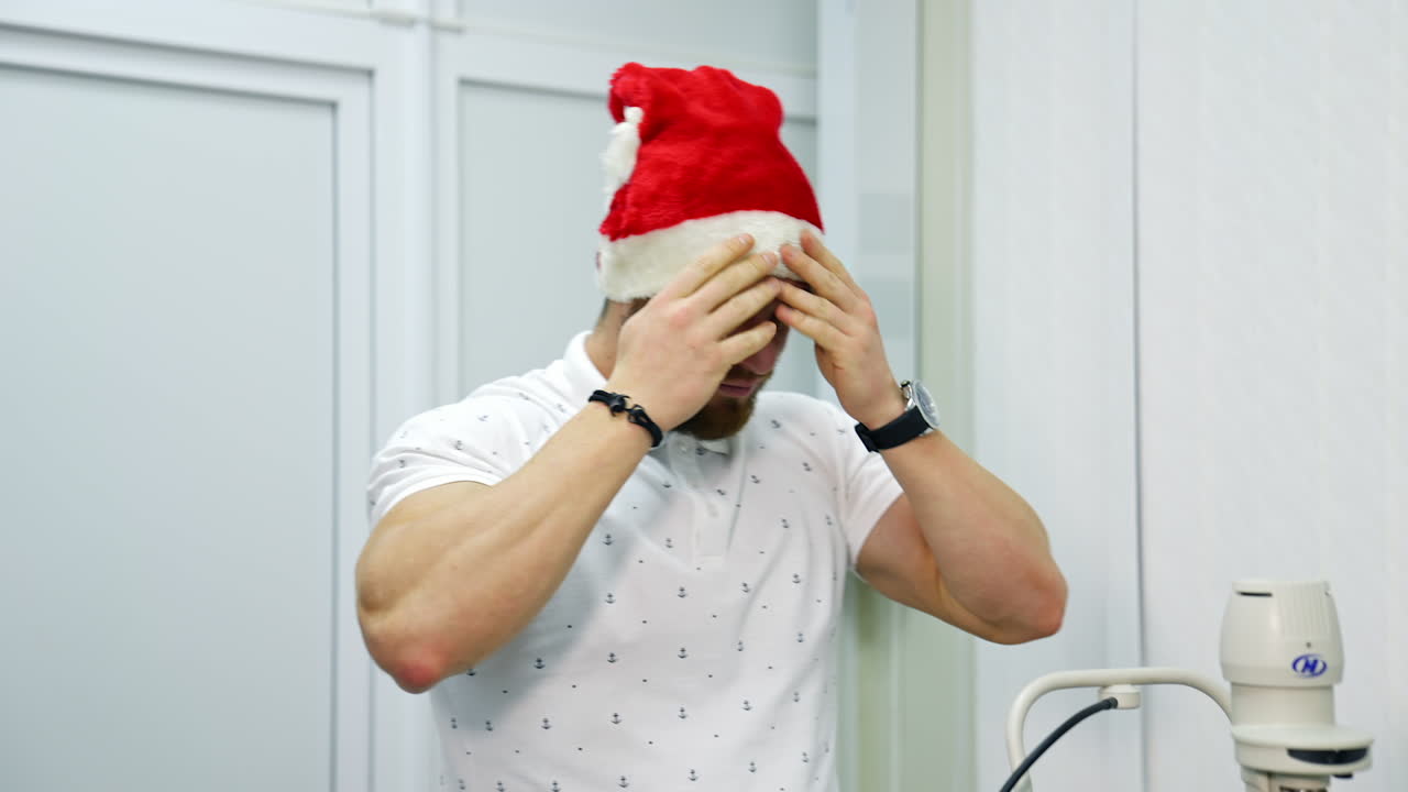 Man putting on Christmas cap in clinic. Smiling man visits ophthalmologist at Christmas. Male patient consulting at oculist.