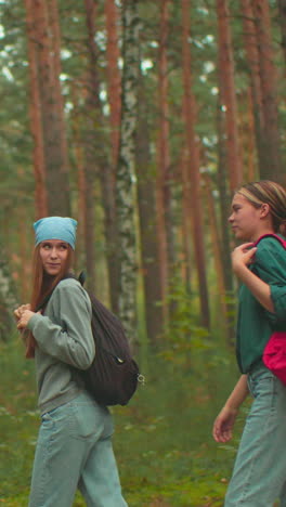 excursionistas de pie en un bosque pacífico, una en una mochila roja ayuda a su amiga en una bufanda azul a ponerse una bolsa negra, comparten una cálida sonrisa mientras ajustan sus bolsas y comienzan a caminar