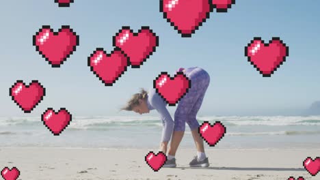 animation of floating red pixel hearts, over woman tying shoe on beach