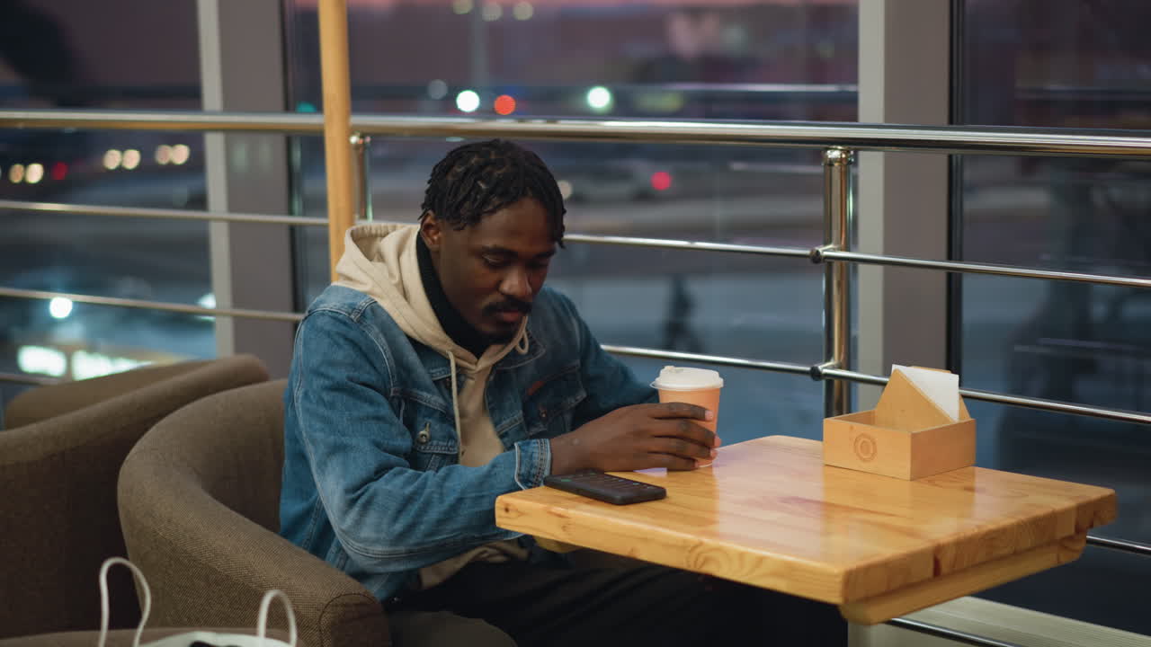 Young man in denim jacket and beige hoodie sits in cozy cafe sipping coffee from takeaway cup while watching traffic through large window, surrounded by warm light, relaxed atmosphere