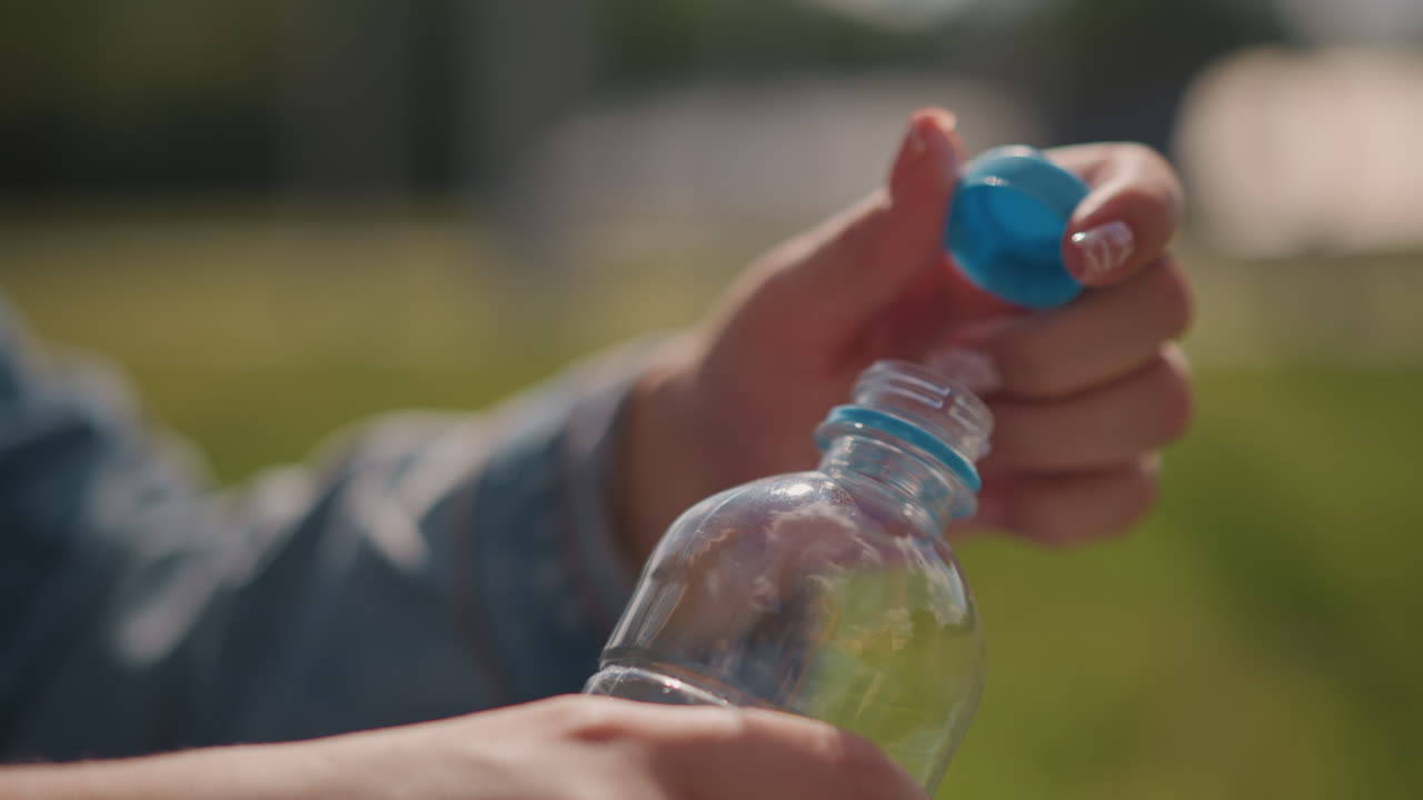 Sormet kiertävät pullonkorkkia, valkoihoinen nainen nurmikolla tauolla, nopea energinen liike farkkuhihan kanssa, tiukka ote ja kuvioitu korkki, nesteytyshetki aktiviteetin välissä, kesävalo ja urheilullinen tunnelma.