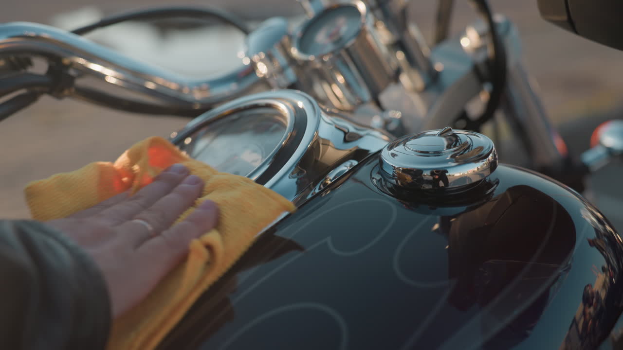Close up of rider hand wiping shiny motorcycle tank with yellow cloth, sunlight reflecting on chrome handlebars and gauges while polishing metal surface for maintenance and care