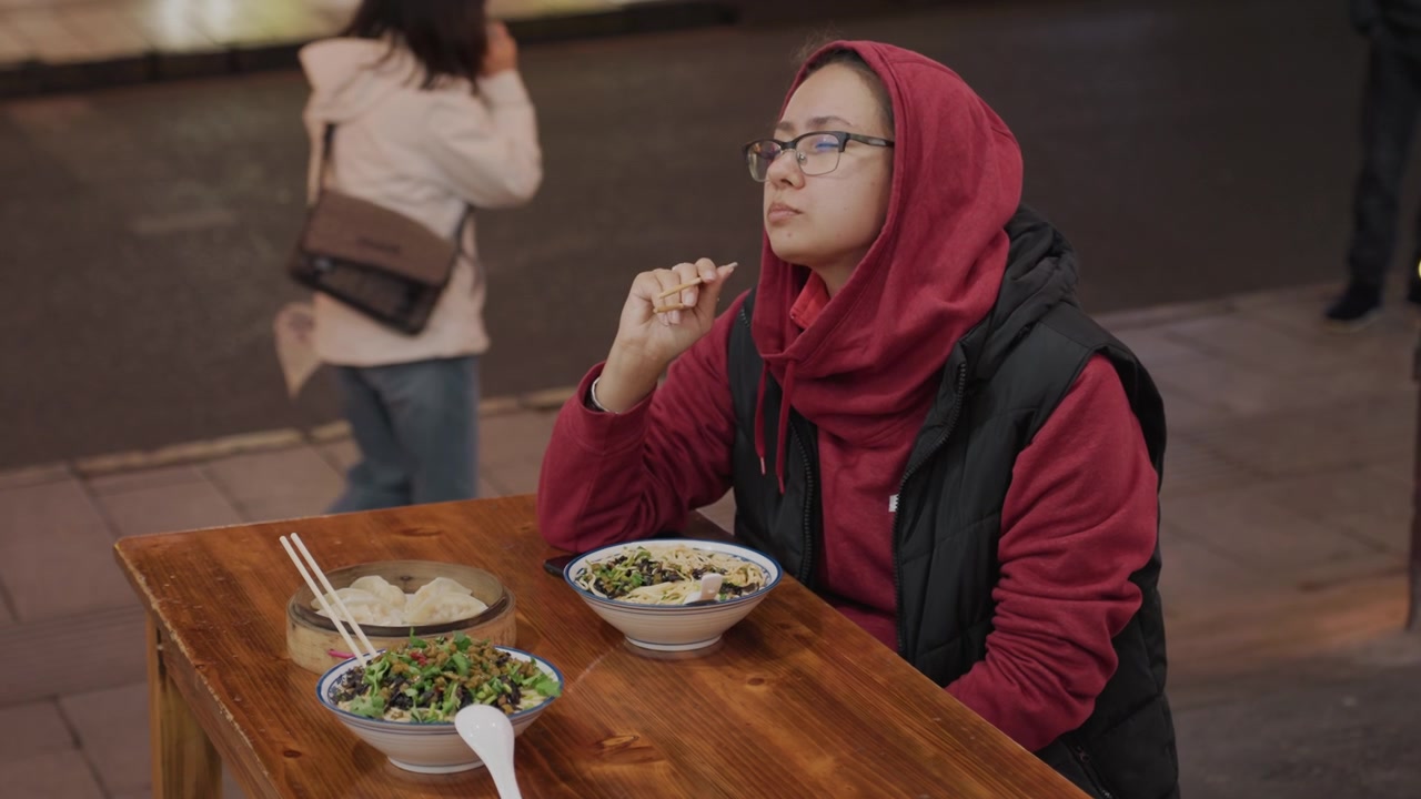 Night street table solo traveler eating, woman in red hoodie pauses from walk to eat noodles at wooden table with chopsticks, ambient traffic lights and bokeh, casual late evening market atmosphere