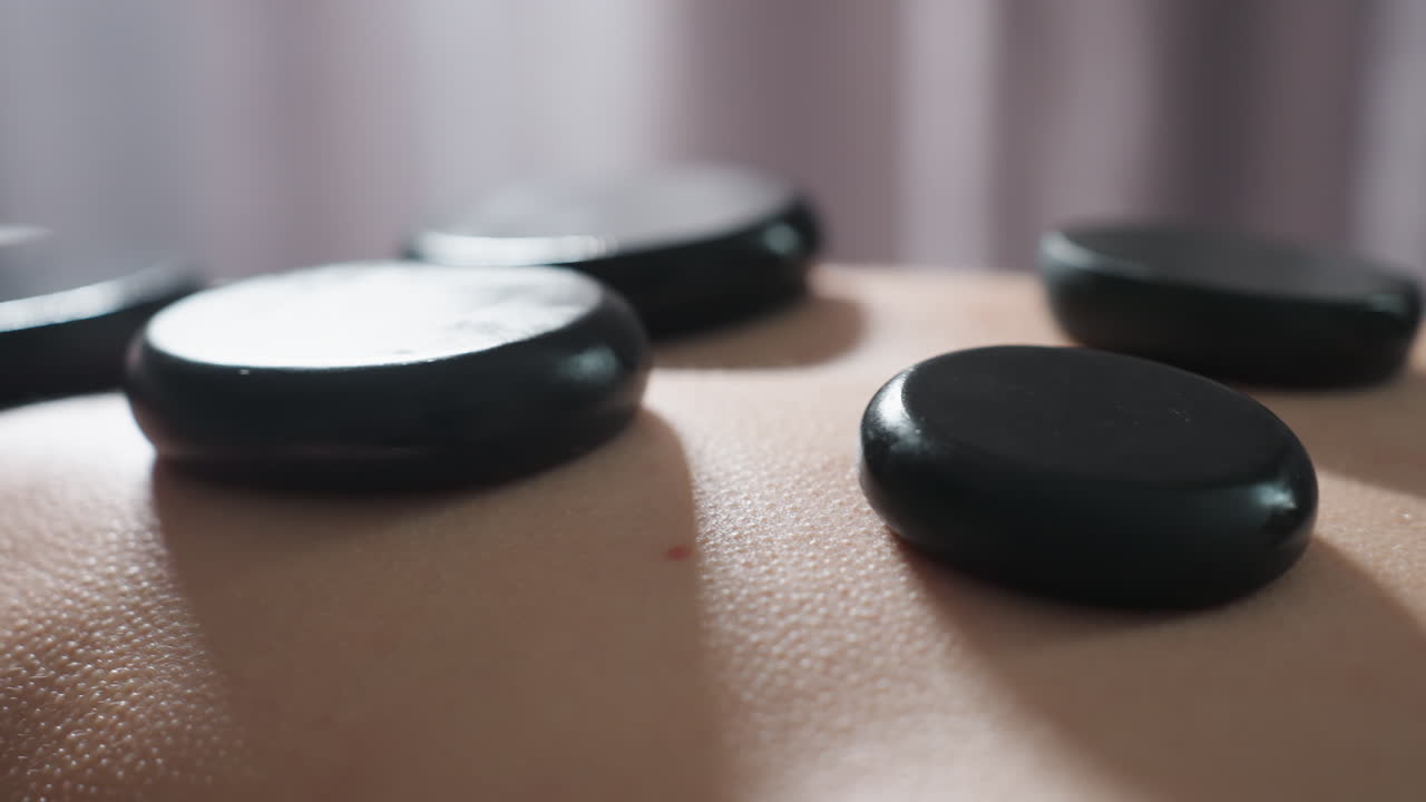 Very close up view of smooth black massage stones placed on client back during spa treatment, highlighting skin texture, warmth, and serenity in soft light setting for deep relaxation
