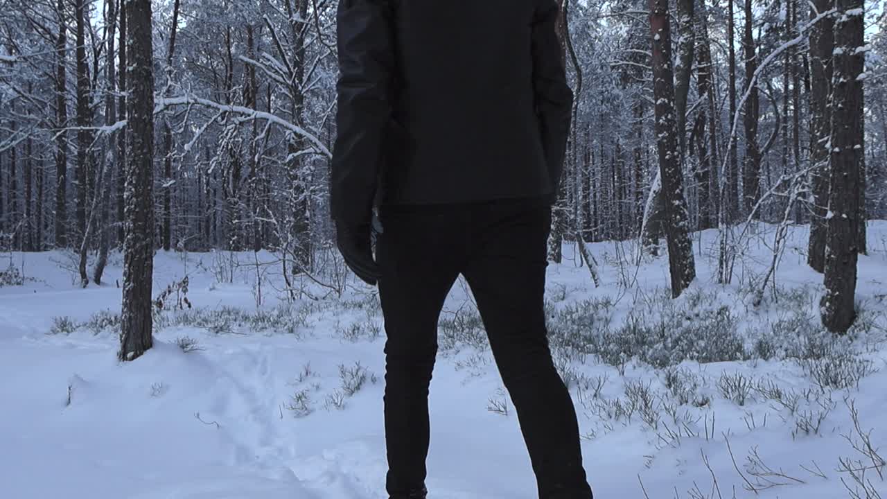Man with black clothing and a black hat walking away from the footage into the winter woods in front of him while the white snow is thick and fluffy. He makes snow fly around with him walking and move