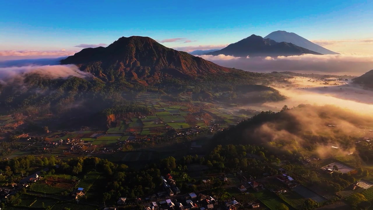 Una mozzafiato rivelazione cinematografica aerea con drone che cattura le maestose silhouette del Monte Batur e del Monte Agung che emergono sopra un mare di nuvole durante un'alba tropicale splendente.