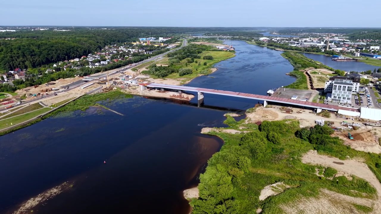 Splendido video con drone cattura il Ponte della Confluenza che attraversa il fiume Nemunas a Kaunas, Lituania. Ampia inquadratura panoramica mostra rive verdi lussureggianti, acqua blu, edifici urbani e cieli estivi sereni