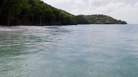 Drone-Wide-shot-of-water-crashing-on-the-beach-shore-sand-buildings-on-hill-in-background-blue-sky-white-clouds-turquoise-water-relaxation-vacation-tourism