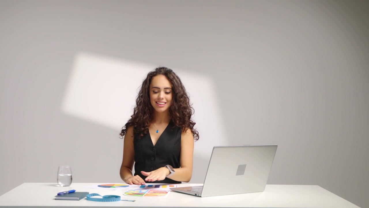 Nutricionista sonriente sentada en el escritorio trabajando en su portátil, haciendo clic y analizando tablas y gráficos de dietas mientras revisa notas y planifica un programa de nutrición saludable en una moderna oficina estudio