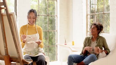 female artist painting on canvas while friend enjoys coffee and conversation at home