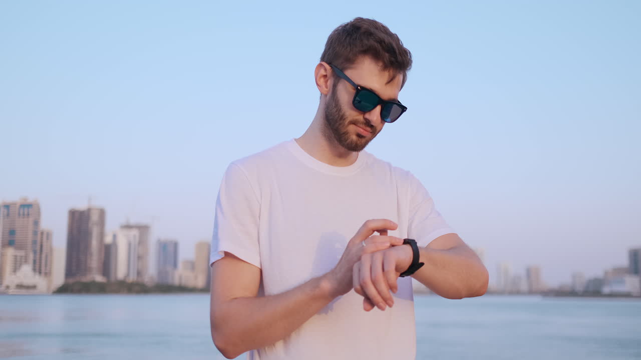 feliz hombre guapo con gafas de sol y camiseta blanca usa un reloj mira y presiona su dedo en la pantalla de pie en la orilla en verano contra la ciudad y los edificios
