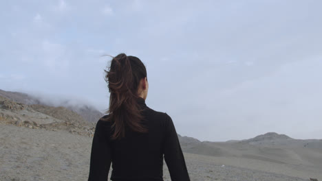 Bella-Mujer-Por-Detrás-Vestida-De-Negro-Parada-En-El-Desierto-Nublado-Por-La-Tarde-Mientras-El-Viento-Sopla-Y-Mueve-Su-Cabello