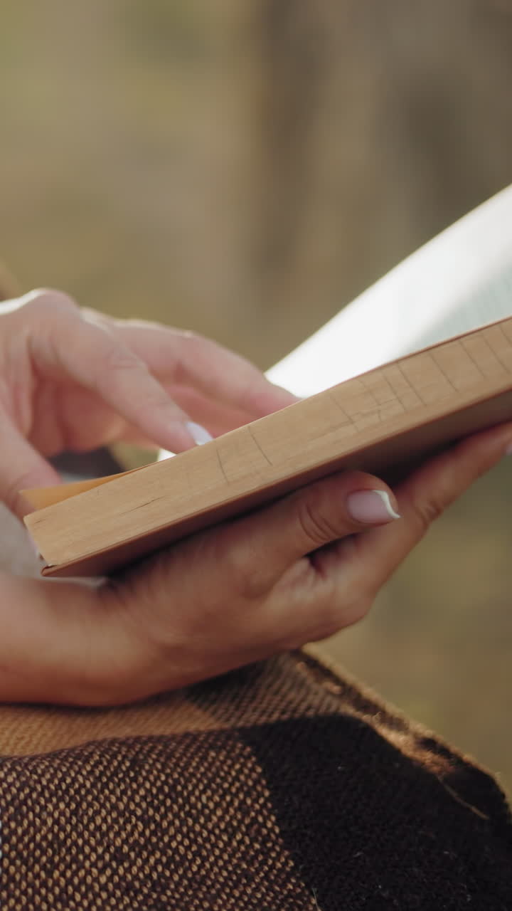 Close-up of a lady hand with a bangle turning to a new page on a checkered blanket, enveloped in a golden glow that highlights the serene outdoor setting
