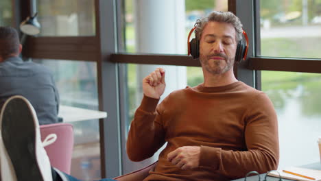 Mature-Businessman-With-Feet-On-Desk-In-Office-Listens-To-Music-On-Wireless-Headphones-Air-Drumming